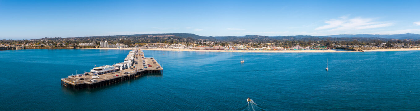 Aerial panorama of the Santa Cruz wharf extending into Monterey Bay with the sandy beach, coastal boardwalk, and hills in the distance on a clear sunny day in California.