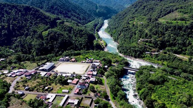 Aerial view of a small settlement with buildings, roads, a sports field and bridge beside a turquoise river in a steep green mountain valley. The scene highlights rural infrastructure, transport and c
