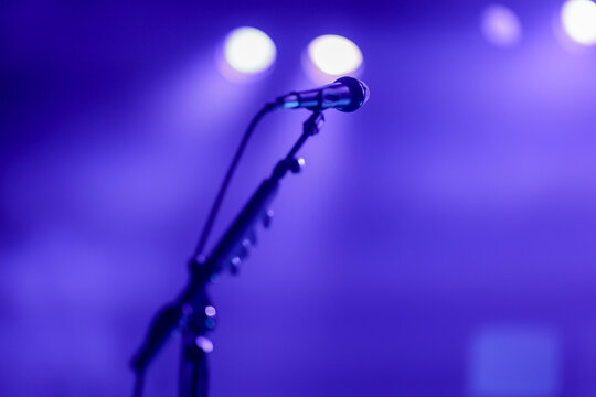 A microphone stands ready under dramatic blue stage lighting, in anticipation of a live concert performance by a singer singing in the mic.