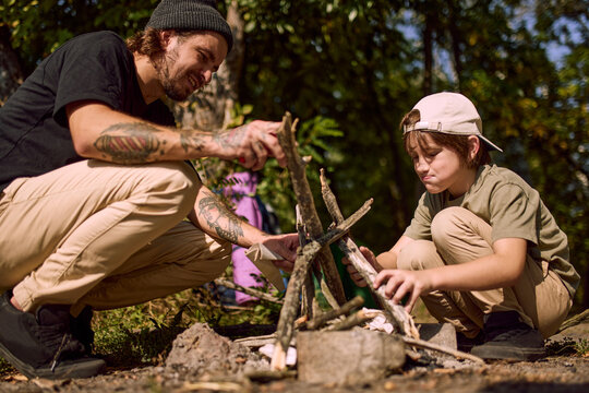Smiling dad and boy build campfire together near lake at forest campsite. Family bonding, outdoor cooking and camping tradition concept.