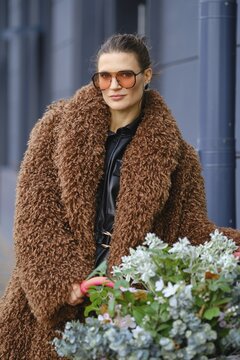 A beautiful woman stands outdoors in a city, wearing a large brown fluffy coat and orange sunglasses. She is posing near a pot of green plants, showcasing her unique sense of style and love for nature