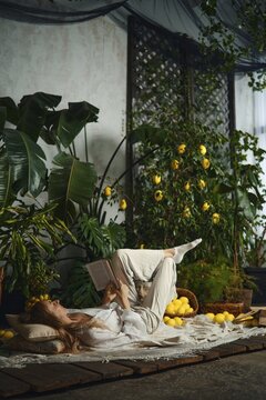 A woman lies on her back with a book in her hands in a room filled with green plants and yellow lemons. She appears relaxed and engaged in reading. The setting is bright and inviting
