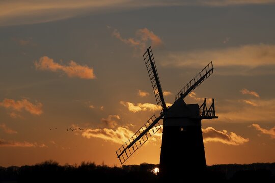 Windmill silhouetted at sunset with a red sky and clouds and a skein or flock of Pink-footed geese (Anser brachyrhynchus) flying above, Burnham Ovary Staithe, Norfolk, England, United Kingdom