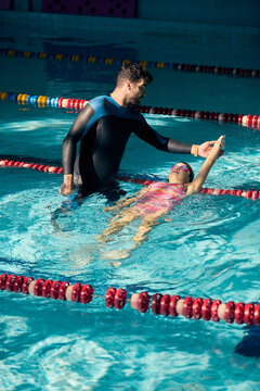 Caring instructor supports little girl floating on back in pool with calm trust. Youth sports, water confidence, active childhood concept.