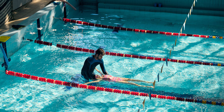 Male instructor supports young girl floating on her back in pool with calm trust. Water confidence, swimming basics, child safety concept.