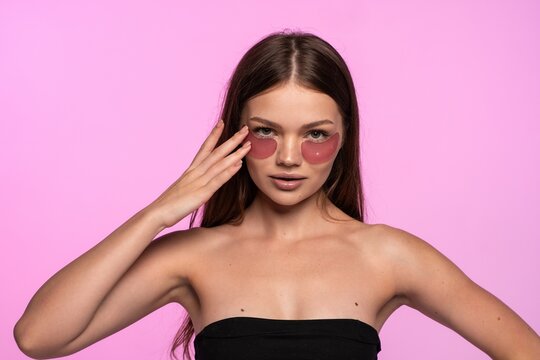 Young woman applying pink under eye patches for skin hydration and wellness, looking directly at camera