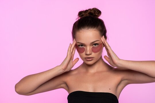 Woman wearing pink eye patch treatment for under eye skin care regime, touching temples on pink background