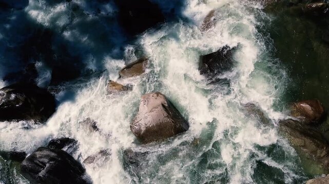 Vertical drone view of powerful whitewater rapids surging around giant boulders. Foaming water and dark shadows create a dramatic scene of natural force, freshwater wilderness and outdoor adventure.