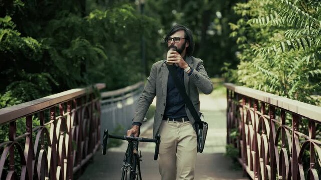 Commuter Carrying Bicycle Walking Across Bridge In City Park Holding Coffee Cup. Modern Active Lifestyle View