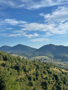 Scenic summer view of a green mountain valley with scattered village houses nestled among rolling hills and dense forests under a bright blue sky with white clouds