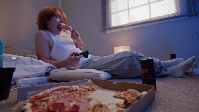 Caucasian man playing video game with controller while eating pizza on floor. Visuals ideal for youth lifestyle campaigns, junk food delivery promos, gaming culture ads