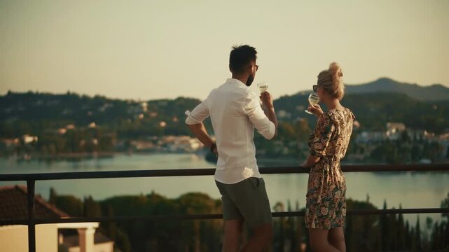 Couple Drinking Wine Glasses Standing On Resort Balcony Terrace Looking At Sea Coast View During Sunset. Evening Romance 