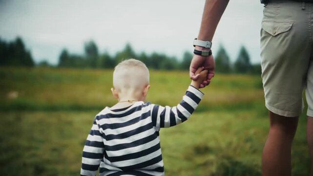 Parent Holding Little Child Hand Walking Together In Green Meadow Field. Toddler Learning To Walk With Adult In Nature