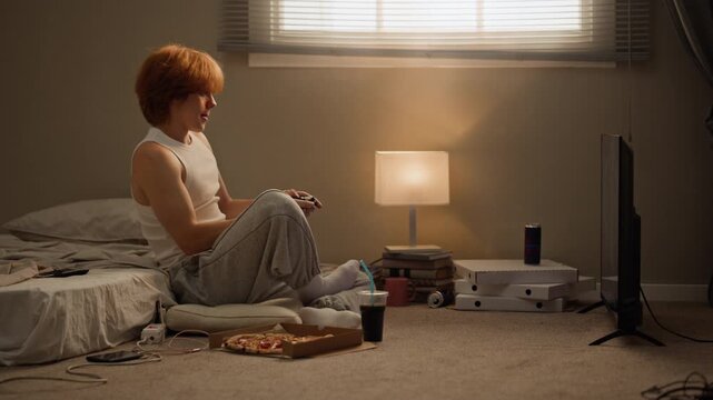 Caucasian man playing video games with controller and eating pizza slice while sitting on floor. Clip ideal for youth leisure, online gaming, fast food delivery, and entertainment ads