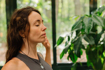 young woman practices yoga and meditation in a room with large windows, with a forest in the background, in the summer