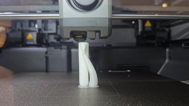 Close-up of a 3D printer print head nozzle extruding white filament layer by layer onto a heated build plate in a workshop.