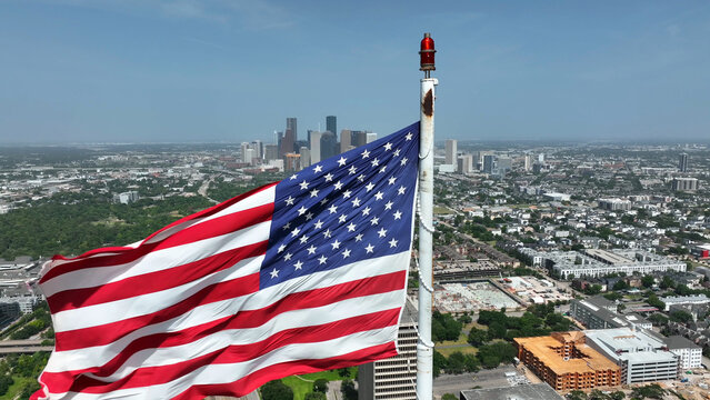 Aerial view of an American flag waving in the foreground with the Houston skyline visible in the background under a clear blue sky in Houston, Texas, United States.