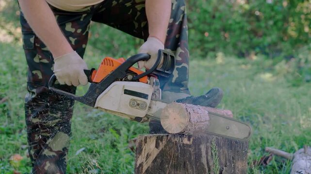 Gloved worker cuts a small log with a chainsaw on a tree stump in a grassy woodland setting, showing active outdoor firewood preparation