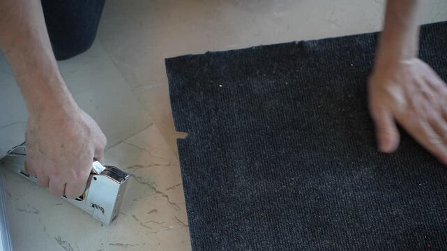 The flooring specialist is securing the carpet to the wooden floor using a stapler.
