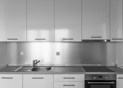 A modern minimalist kitchen features glossy white cabinets, a stainless steel sink, and an embedded stovetop with an oven beneath.