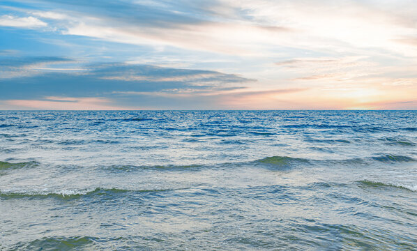 A serene calm sea featuring gentle rippling waves under a vast blue sky with soft evening clouds at sunset.