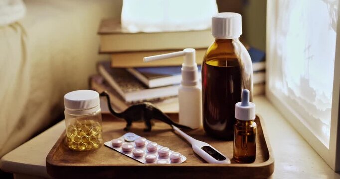 Pediatric medicine and healthcare items on a wooden tray at a child's bedside, featuring cough syrup, pills, digital thermometer, and a toy dinosaur for home illness care.