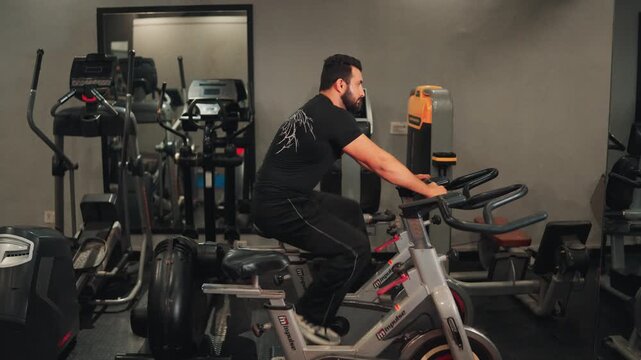 Side profile view video shot of an athletic man riding a stationary exercise spin bike during an indoor cardio cycling workout in a modern gym.