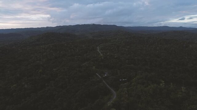 Wide aerial view of a remote road winding through an immense tropical rainforest landscape beneath heavy clouds. Small roadside buildings emphasise the scale and isolation of the forested region.