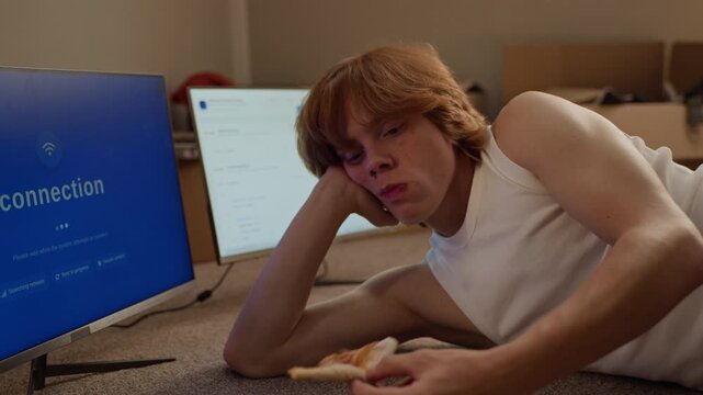 Caucasian young man lying on floor eating pizza and typing on keyboard before monitors. Footage suitable for IT startups, software development, coding bootcamps and student lifestyle videos