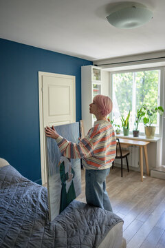 Focused woman holding large canvas with painting standing beside bed in bedroom, positive female looking at wall and planning visualizing where to hang own artwork, creative home organization.