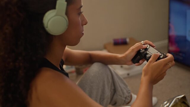 Young adult multiethnic woman wearing headphones playing video game with wireless controller on television. Ideal for esports commercials, gaming technology ads, and entertainment campaigns