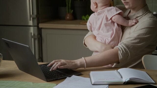 Caucasian young adult woman holding infant baby while working remotely on laptop at home table. Footage suitable for work life balance, maternity leave and freelance career campaigns