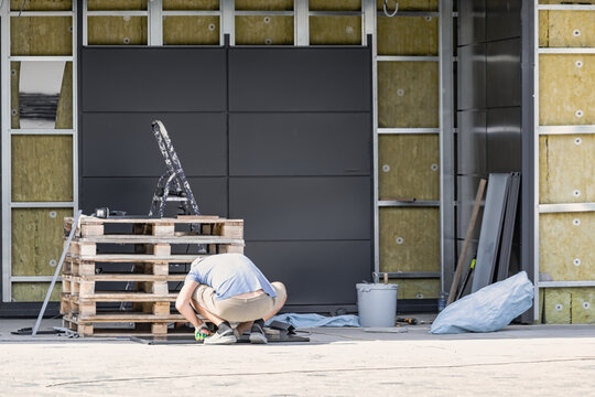A worker is crouched down, inspecting materials on wooden pallets at a construction site. Tools and a bucket are nearby as work continues on the building