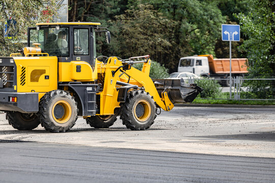 A construction loader moves material along a road where work is underway. The scene depicts activity in an urban environment with nearby vehicles