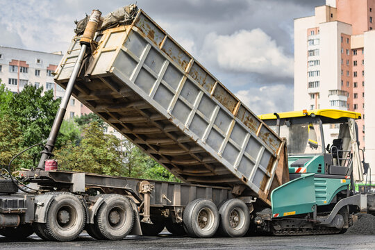 A dump truck lifts its body to unload asphalt into a paving machine at a construction site. Asphalt paving and road surface modernization