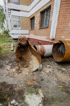 Rusted metal pipes rest on the ground next to a brick structure. Parts of the walls show damage. Overgrown grass surrounds the area. It is daylight