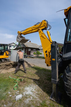 A worker is using a backhoe to dig near a building. Another machine is parked nearby. The sun is shining and the scene shows construction activity