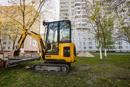 An excavator is parked on the grass in front of a residential building. Trees surround the area while the machine prepares for work on the ground
