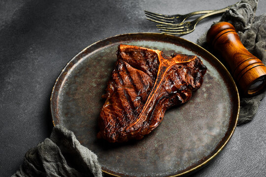 Juicy grilled T-bone steak served on a dark plate. Delicious beef with perfect grill marks offers rich flavor. Great background for steakhouse menu design presentation.