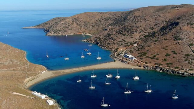 Aerial view of a double-sided bay with sailboats anchored in clear blue water, surrounded by rocky hills and coastal terrain in Islands, Greece.