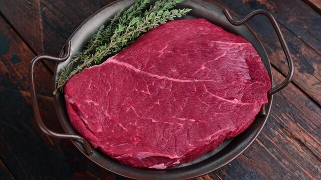 Close-up of raw lean beef steak with fresh thyme in a dark handled metal dish rotating over a rustic brown wooden tabletop.