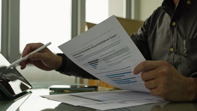 Investor holding financial research report document at office desk beside tablet computer, analyzing shareholder distribution data, market recommendation spread, equity metrics.