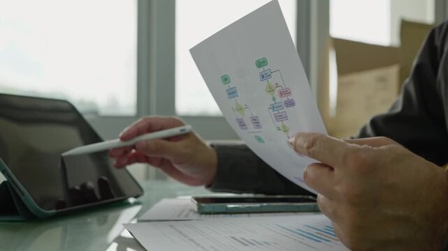 Businessman analyzing flowchart diagram paper sheet at glass desk beside digital tablet smartphone financial charts in bright office.