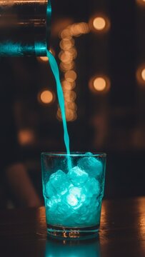 Vibrant blue cocktail being poured from a shaker into a glass filled with ice cubes in a moody bar setting with glowing bokeh lights