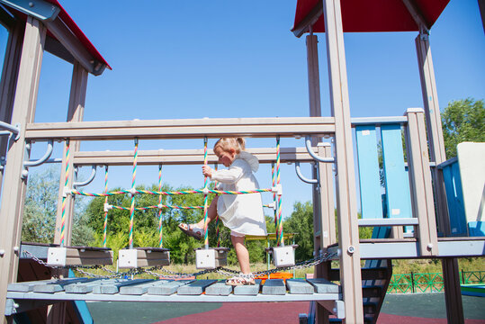 A girl in a white dress plays on the playground, climbs a rope ladder, and on the bars at the top of the slide.