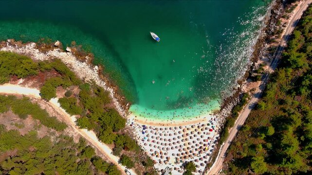 Aerial view of a small boat floating in the turquoise waters of a beach in Thasos with rows of sun umbrellas on the sand in Thasos, Greece.