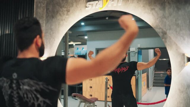 Reflection in a large arch mirror of a bearded man flexing upper body muscles and checking physique progress in a modern gym.