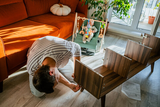 Father building new tv stand furniture on the floor, carefully following instructions while his baby watches from a bouncer in the brightly lit living room, creating a family home