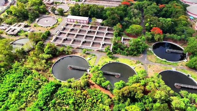 Aerial view of a wastewater treatment plant with circular sedimentation tanks and rectangular aeration basins surrounded by lush green trees in Surat, Gujarat, India.
