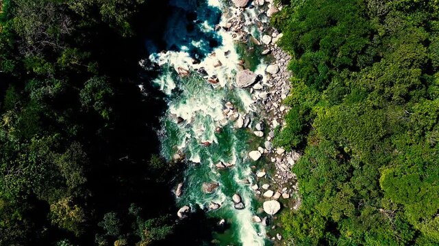 Drone view looking straight down on turquoise rapids rushing among large boulders through dense tropical forest. Contrasting sunlight and shadow reveal the river&rsquo;s dynamic flow and rugged natural sett
